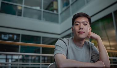 Brian Hie, assistant professor of chemical engineering in the School of Engineering, is wearing a light gray shirt sitting indoors with their head resting on one hand, looking toward the camera.