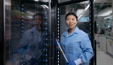 Lingyin Li, professor of biochemistry in the School of Medicine, is wearing a blue lab coat and gloves holding a pipette, standing in front of an open laboratory refrigerator.